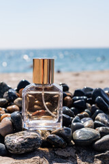 A transparent bottle of perfume with a golden cap stands on a pebble beach, blue water of the sea, the ocean. A blank bottle mockup. vertical view