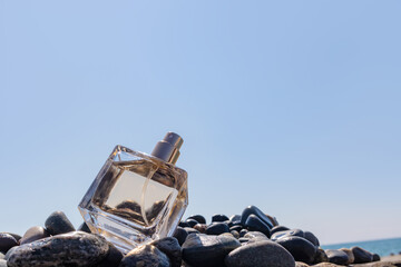 A glass bottle of perfume among the pebble stones against the blue sky. Presentation of the summer fragrance of freshness of perfume. A copy space.