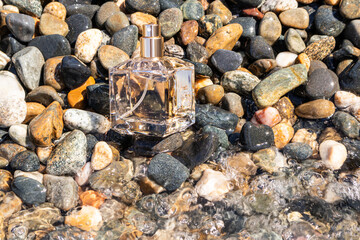 A transparent square bottle of perfume stands among the pebbles washed by sea water. The aroma of a fresh sea breeze. A blank bottle mockup.