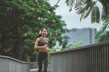 Obraz premium Happy young Asian woman in sportswear jogging and listening to music with headphones on a bridge in a green park for fitness and healthy lifestyle concept