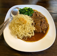 Egg noodles served with braised beef in rich brown gravy, accompanied by blanched green vegetables on a white plate. This image represents Asian comfort food, street food cuisine, and traditional food