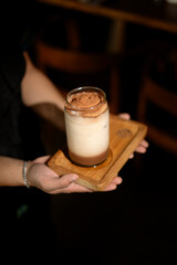 Serving elegant layered coffee drink with foam and cocoa on wooden tray in a dark setting.