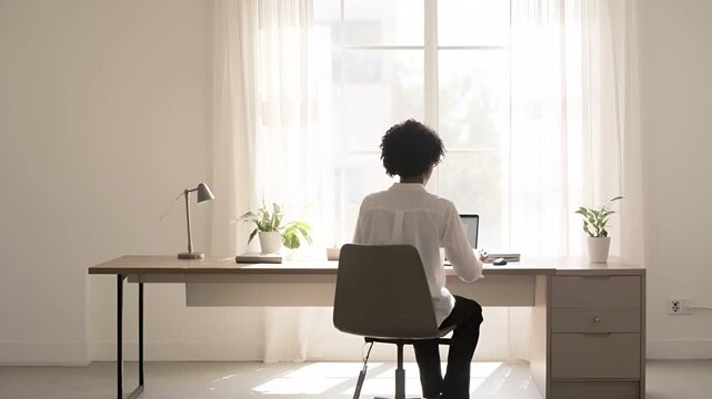 デスクに座りノートパソコンに向かう人物を背後から捉えたワイドショット。
窓から入る柔らかな自然光が室内を均一に照らす。
白や淡いグレーを基調としたミニマルなオフィス空間。