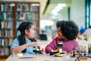 Fototapeta premium African American girl guides robotics experiment alongside Asian girl, problem solving activity. Black girl develops technology skills through shared robotics work, classroom collaboration.