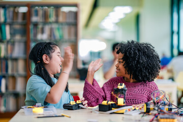 Two children celebrate robotics project success inside library, smiling with raised hands, robotic...