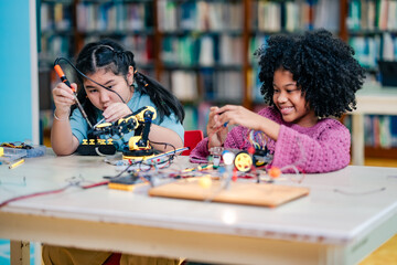African American girl engages robotics task alongside Asian girl, assembling electronic parts, STEM education focus, diversity inclusion, teamwork skill building, creative learning atmosphere.