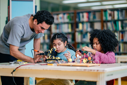 Instructor assists students with robotics project, demonstrating wiring technique, electronic components, educational guidance, STEM learning, collaborative classroom, focused technology instruction. - Powered by Adobe