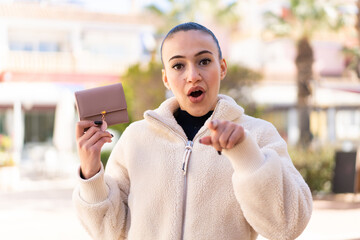 Young moroccan girl holding a wallet at outdoors surprised and pointing front