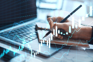 Business person analyzing financial chart and writing notes beside laptop on office desk with abstract digital stock graph overlay concept.