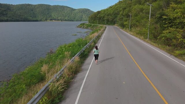 Rear view of an Asian woman jogging alone on a quiet road beside a lake, surrounded by green hills and natural outdoor scenery. Healthy lifestyle