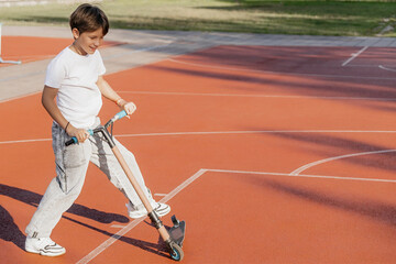 Smiling young boy riding a scooter on an empty outdoor basketball court on a sunny summer day, enjoying physical activity and childhood freedom. Copy space