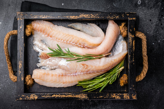 Ready for cooking Raw Hake fish fillets in a wooden tray with herbs. black background. top view