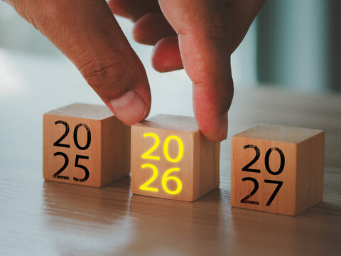Wooden calendar blocks showing transition from 2025 to glowing 2026 with hand adjusting middle cube, hopeful new year concept and optimistic mood