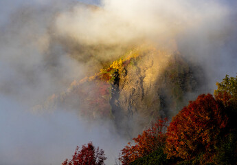 Fog covers a rocky mountain as bright sunlight touches the top. Trees in red and orange surround the base, showing fall colors in the early morning.