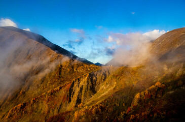 Clouds roll over the mountain peaks and tiny houses in a valley in the sunset light. Trees line the slopes with colors changing for the season. Nature shows its beauty in this mountain region.