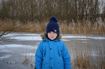 Child boy standing  in winter coat with snowy background near river in residential area.