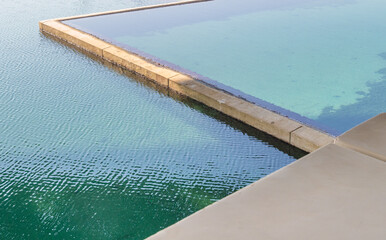 Water fills a pool, creating a clear line where it meets the edge. The sun shines down, showing ripples on the surface and a clear, blue sky above.