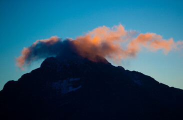 A mountain stands tall as clouds gather around its peak during sunset. The colors in the sky change, marking the end of the day with a natural display.