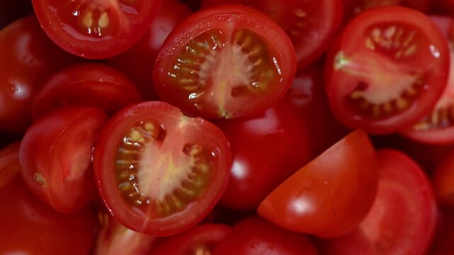 A vibrant seamless 4K video loop of fresh cherry tomatoes, cut in half, rotating smoothly on a dark surface, filmed from a top-down perspective. The clip highlights the bright red color, glossy textur