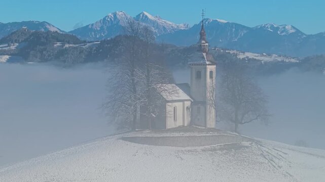 Breathtaking Winter Mountain Range. iconic Jamnik church in Slovenia Julian Alps in the foggy mist. Aerial 4k 60fps drone footage