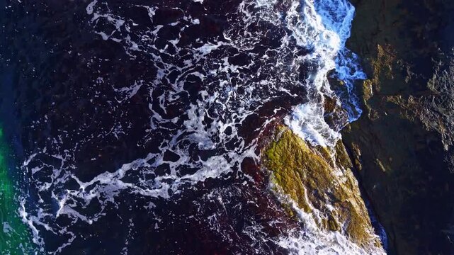 Bright morning light illuminates the contrasting colors of the rocky shore as waves rhythmically crash, blending deep blues with striking greens and earthy tones in a coastal spectacle.