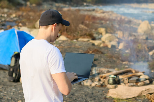 Man typing on laptop from behind at campsite with fire and river backdrop.
