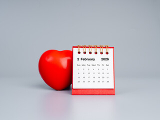 Minimal desk calendar showing February 2026 with a red heart ball beside it on a gray background. Symbolizes the month of love and is ideal for Valentine&rsquo;s Day visuals and romantic themes.