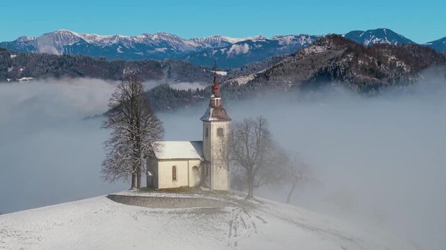 Cinematic scenery of winter foggy sunrise, drone orbit flight around iconic Jamnik church in Slovenia, covered by snow and myst. aerial 4k 60 fps footage