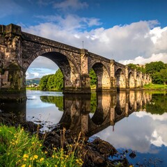 Fototapeta premium Historic Stone Bridge Reflected in Calm River Waters Under a Blue Sky.