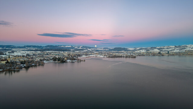 Aerial drone view of Cham on Lake Zug in winter, where a snow-covered landscape meets calm water at dusk, creating a quiet and atmospheric evening scene.