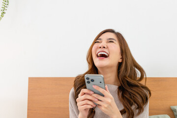 Happy Asian woman laughing at funny content on smartphone in bed