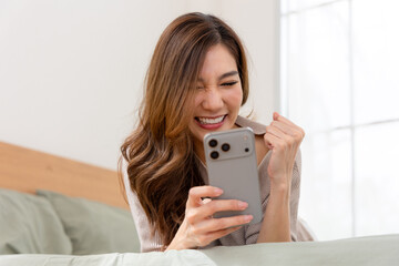 Excited Asian woman celebrating success with smartphone in bed