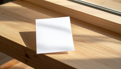 Empty square white paper note card casting a distinct shadow on a warm, sunlit light wooden surface, offering a clean and minimalist backdrop for design or text