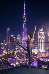 Flexible woman doing ballet pose on balcony with night city skyline view. Urban fitness concept with illuminated skyscrapers and purple lights