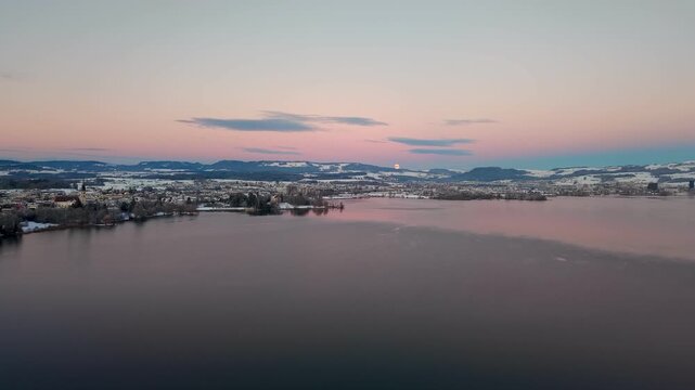 Aerial drone view of Cham on Lake Zug in winter, where a snow-covered landscape meets calm water at dusk, creating a quiet and atmospheric evening scene.