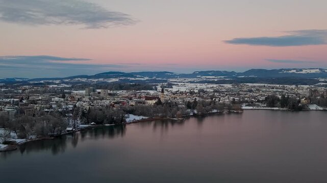 Aerial drone view of Cham on Lake Zug in winter, where a snow-covered landscape meets calm water at dusk, creating a quiet and atmospheric evening scene.