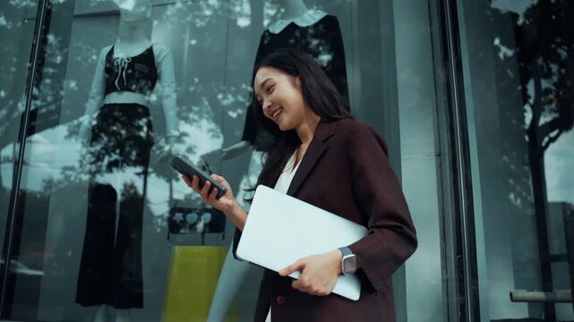 Happy asian professional woman using her phone while standing in front of a shop window
