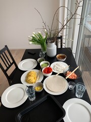 Asian dinner table with fresh ingredients prepared for home cooking; modern home dining table prepared for an Asian meal, featuring rice paper sheets, rice noodles, shrimp, vegetables, herbs, sauces