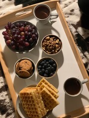 Cozy breakfast tray with waffles, fruit, nuts and tea; a cozy breakfast served on a wooden tray, featuring golden waffles, fresh grapes and blueberries, walnuts, cookies, and cups of hot tea