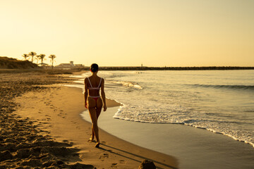Athletic woman walking alone along tropical beach at sunrise, wearing red bikini, seen from behind, casting long shadow on sand, palm trees and golden morning light in the background