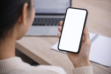 A woman in a cream sweater holds a smartphone with a blank white screen while seated at a desk with a laptop and notebook, viewed over her shoulder in a modern workspace