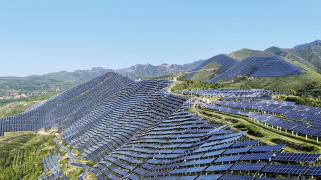 Aerial video of solar panels covering mountain slopes, forming a vast renewable energy landscape under clear blue sky.