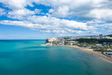 Obraz premium Wide aerial view of Peschici's sweeping sandy bay and turquoise sea, with white buildings clustered on the headland under a sky filled with scattered clouds.