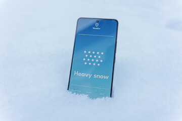 Smartphone standing in snow with heavy snow forecast on weather app screen, winter storm concept