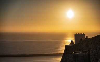 Golden Sunset Over Calm Sea With Silhouetted People On Coastal Wall By A Barrel, Agadir, Morocco