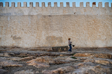 Person With Hat Carrying Bucket Walks Along Historic Stone Wall With Battlements Under Blue Sky, Agadir, Morocco