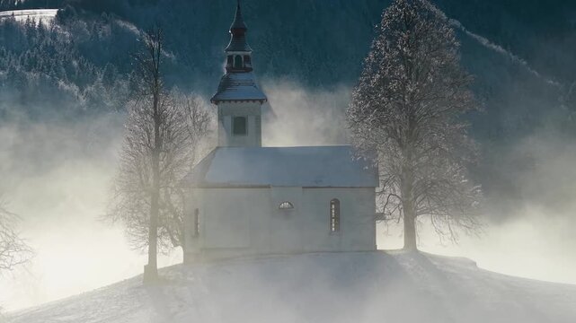 Cinematic landscape of winter foggy sunrise, drone flight approach Jamnik church in Slovenia, covered by snow and myst. aerial 4k 60 fps footage