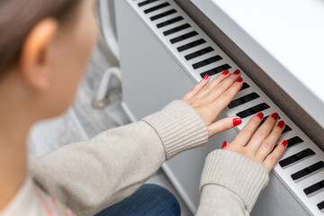  A girl warms her hands by holding them over a radiator. A girl warms herself near a heating radiator. heating radiator. heating season. cold season. 
