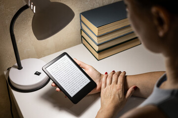 A woman reads an e-reader by the light of a table lamp. A woman reading an e-book at home. reading a book at the table. woman holding e-book. hand holding ereader. 