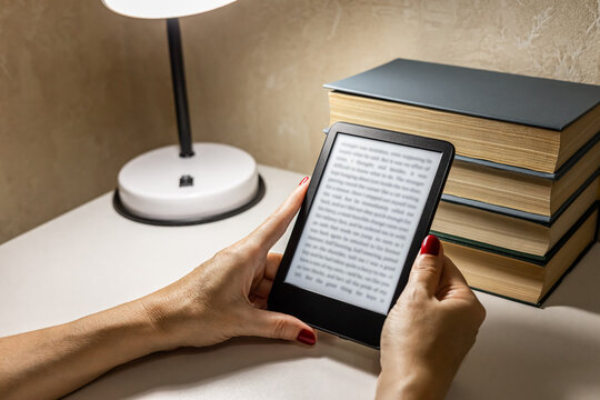 A woman reads an e-reader by the light of a table lamp. A woman reading an e-book at home. reading a book at the table. woman holding e-book. hand holding ereader. e-ink book. 